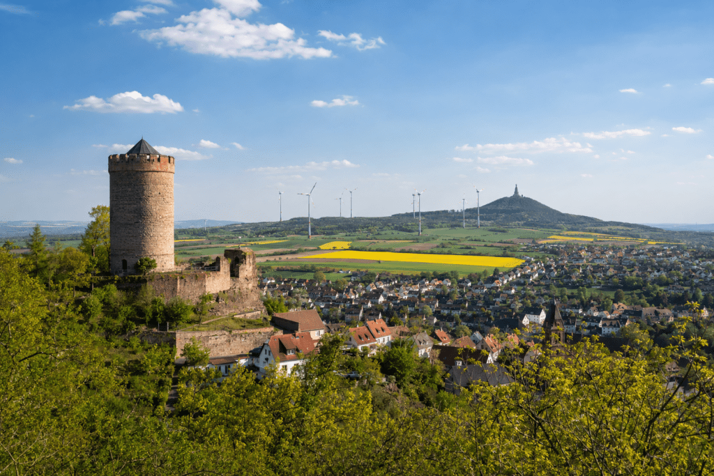 Das Bild zeigt eine eindrucksvolle Panoramaansicht des Kyffhäuserkreises. Im Vordergrund erhebt sich eine historische Burgruine mit markantem Rundturm über einer Stadt mit traditionellen Wohnhäusern und Kirchturm. Die umliegende Landschaft ist geprägt von Feldern, Wäldern und sanften Hügeln, die den ländlichen Charakter der Region unterstreichen. In der Ferne ist das bekannte Kyffhäuser-Denkmal auf dem Kyffhäusergebirge zu erkennen, eines der bedeutendsten Wahrzeichen Thüringens. Die Aufnahme verbindet Geschichte, Natur und Lebensqualität und vermittelt die besondere Attraktivität des Kyffhäuserkreises als Wohn-, Erholungs- und Wirtschaftsstandort. - ✅ WEKEYS IMMOBILIEN – Menschlich. Engagiert. Erfolgreich.